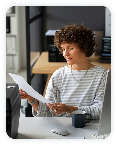 Woman sitting in front of a desktop computer looking at a piece of paper she's holding in her hands.