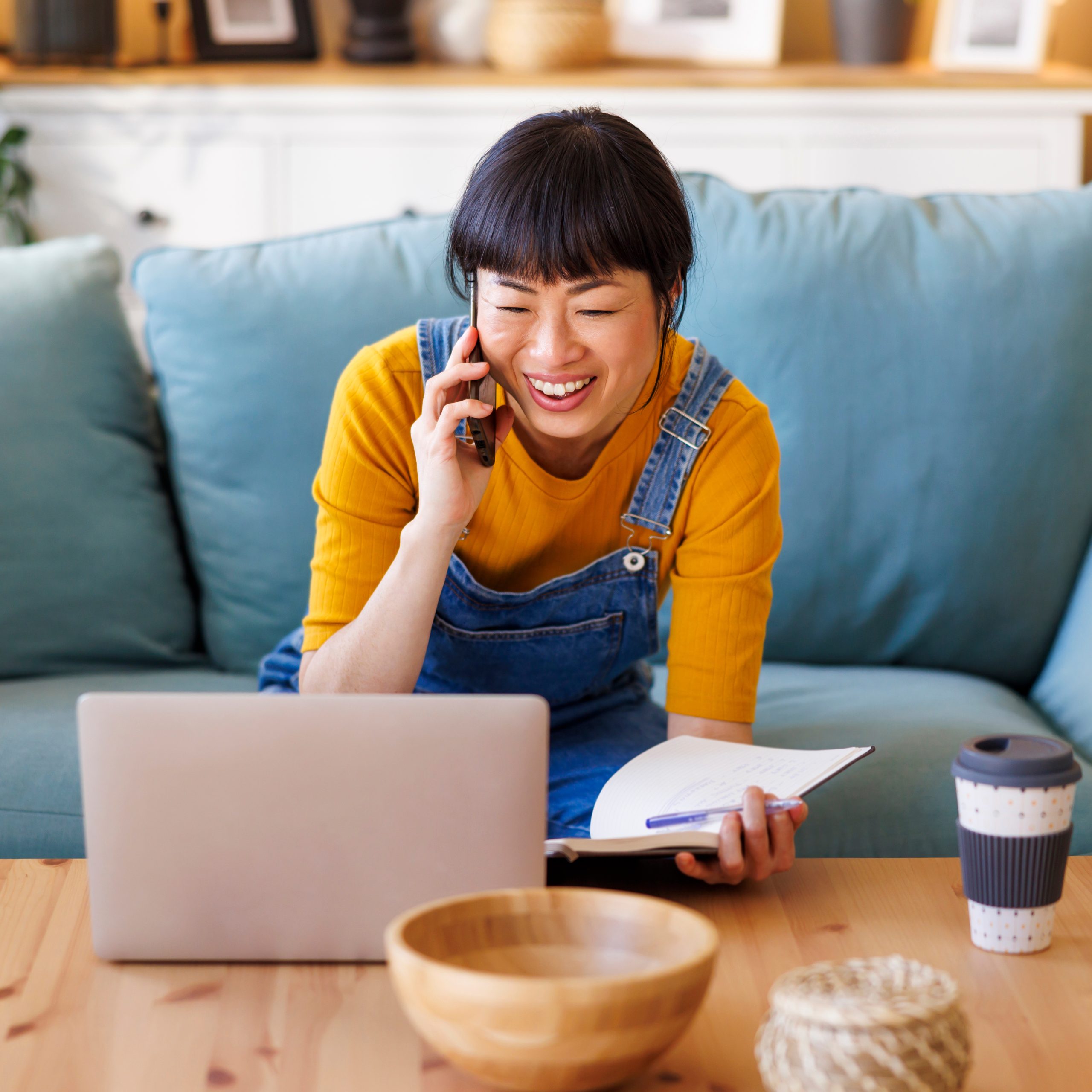 A woman work while seated on the couch, talking on the phone, smiling at her laptop, and holding a notebook