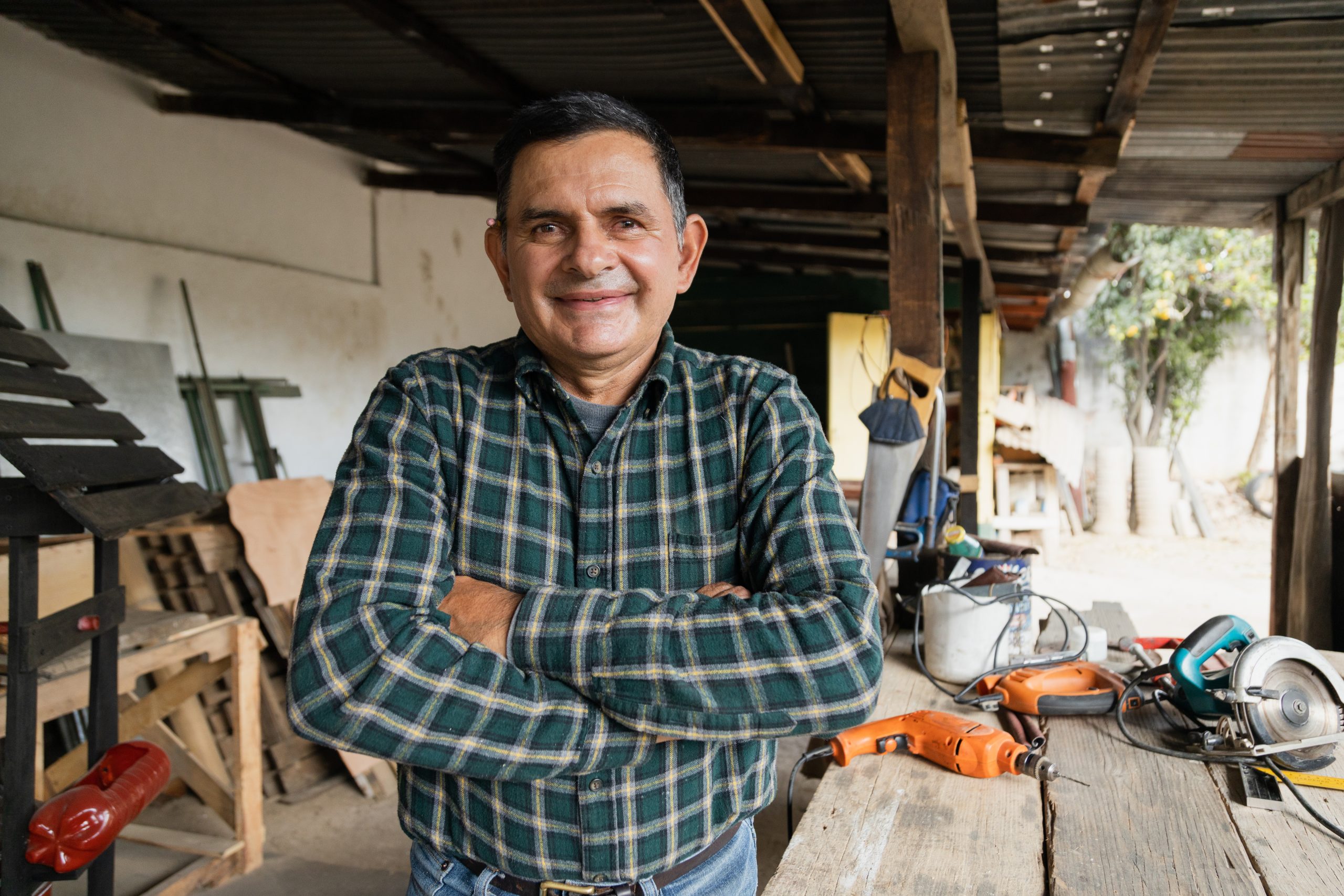 A man in a plaid shirt, smiling with his arms crossed, in front of a work table and tools